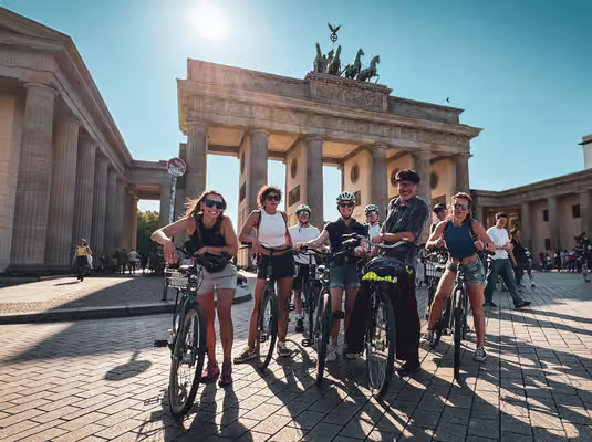 Group of riders gathered at the Brandenburg Gate during a berlin bike tour with sunny skies and historic landmarks in the background.