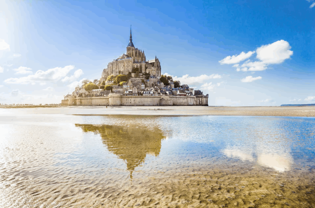 Mont Saint Michel tours featuring the island abbey reflected in shallow tidal waters under a bright blue sky in Normandy.