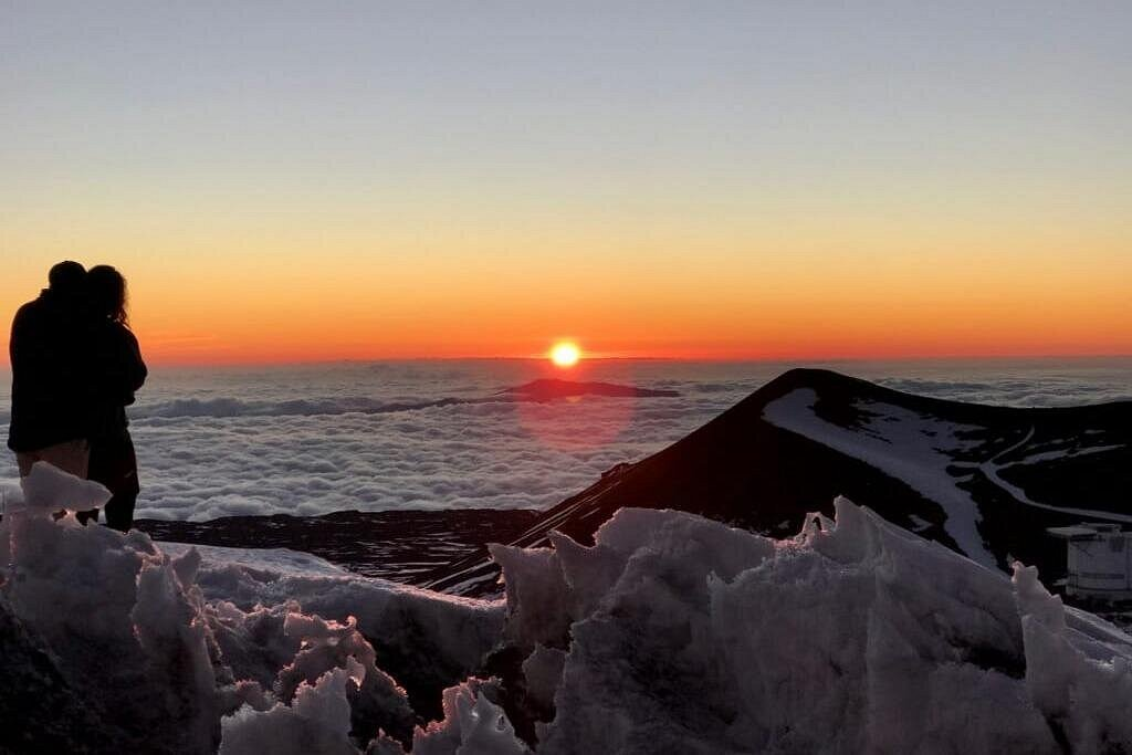 Couple watching sunrise above the clouds on Mauna Kea’s snowy summit during mauna kea stargazing tours, with volcanic peaks and a sea of clouds stretching across Hawaii’s high-altitude landscape