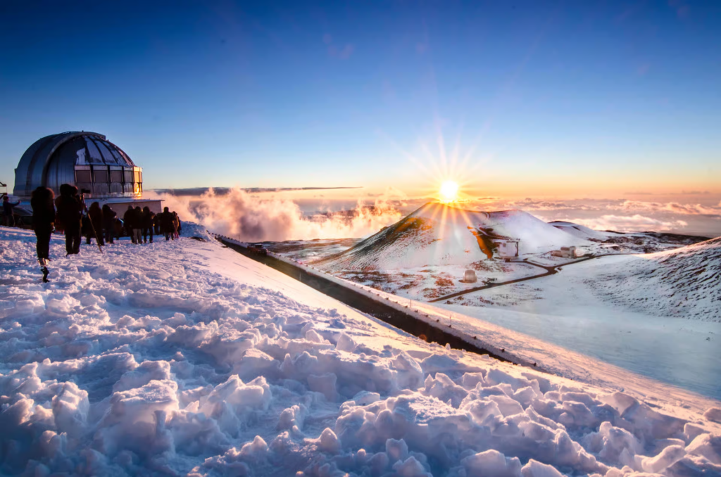 Visitors gather near Mauna Kea’s summit observatory at sunrise during mauna kea stargazing tours, overlooking snow-covered volcanic peaks rising above the clouds in Hawaii