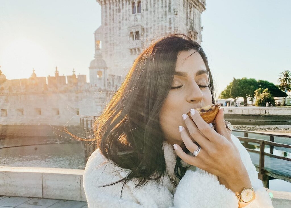 Woman enjoying a traditional pastel de nata pastry at Belém Tower in Lisbon during golden hour, capturing the cultural food experiences often included in lisbon city tours.